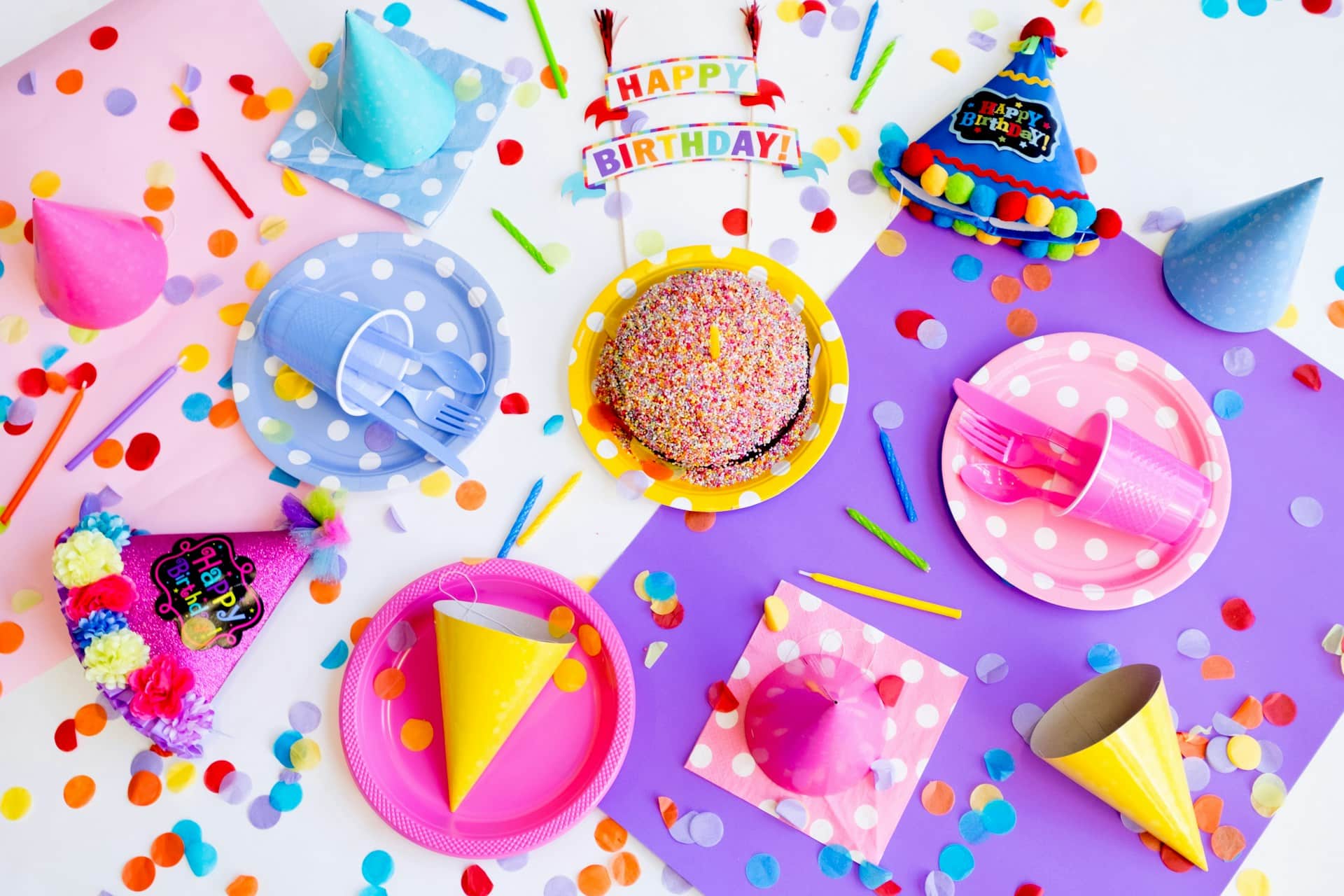 birthday plates and hats in different colors with confetti spread around and a kake in the middle